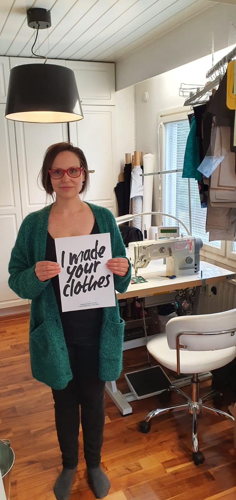 Keikku's founder in her workshop in Finland, holding a hand-lettered sign reading “I made your clothes”, with an industrial sewing machine and garments on a rail behind her.