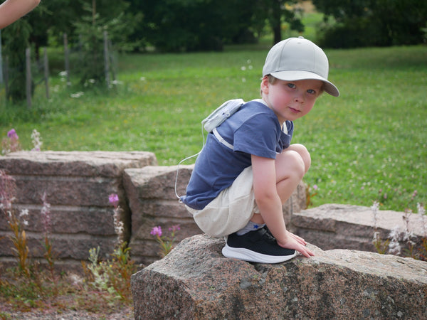 A young child crouching on a stone wall in a summer meadow, wearing a pale blue Keikku mini backpack that holds his insulin pump.