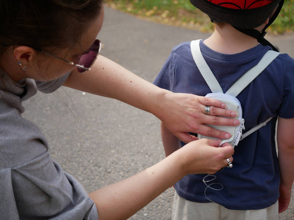 An adult adjusting a pale blue Keikku mini backpack on a young child’s back outdoors, with the insulin pump tubing running from the pouch.