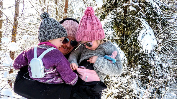 An adult carrying two small children through a snowy forest; one child wears a pale blue Keikku pump backpack, the other holds a pink Keikku pouch.