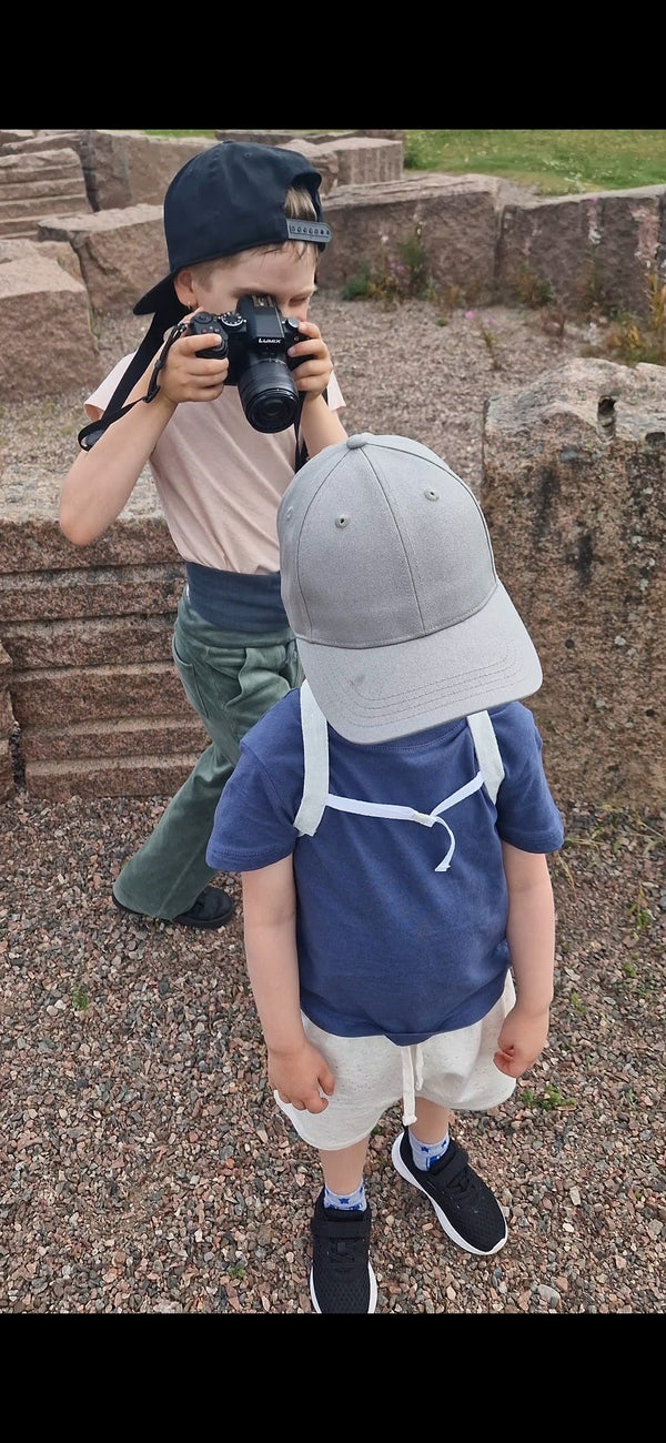 Two children outdoors on a gravel path; the younger one wears a pale blue Keikku mini backpack carrying his insulin pump while an older child photographs him.